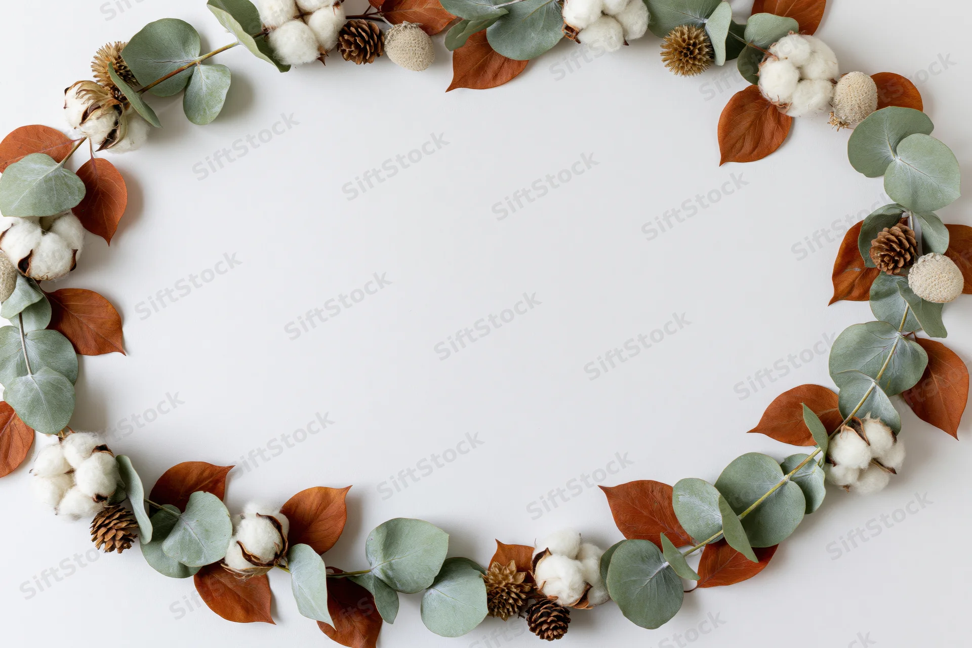 Circular wreath of eucalyptus leaves, dried cotton bolls, and pinecones arranged on white surface with central copyspace