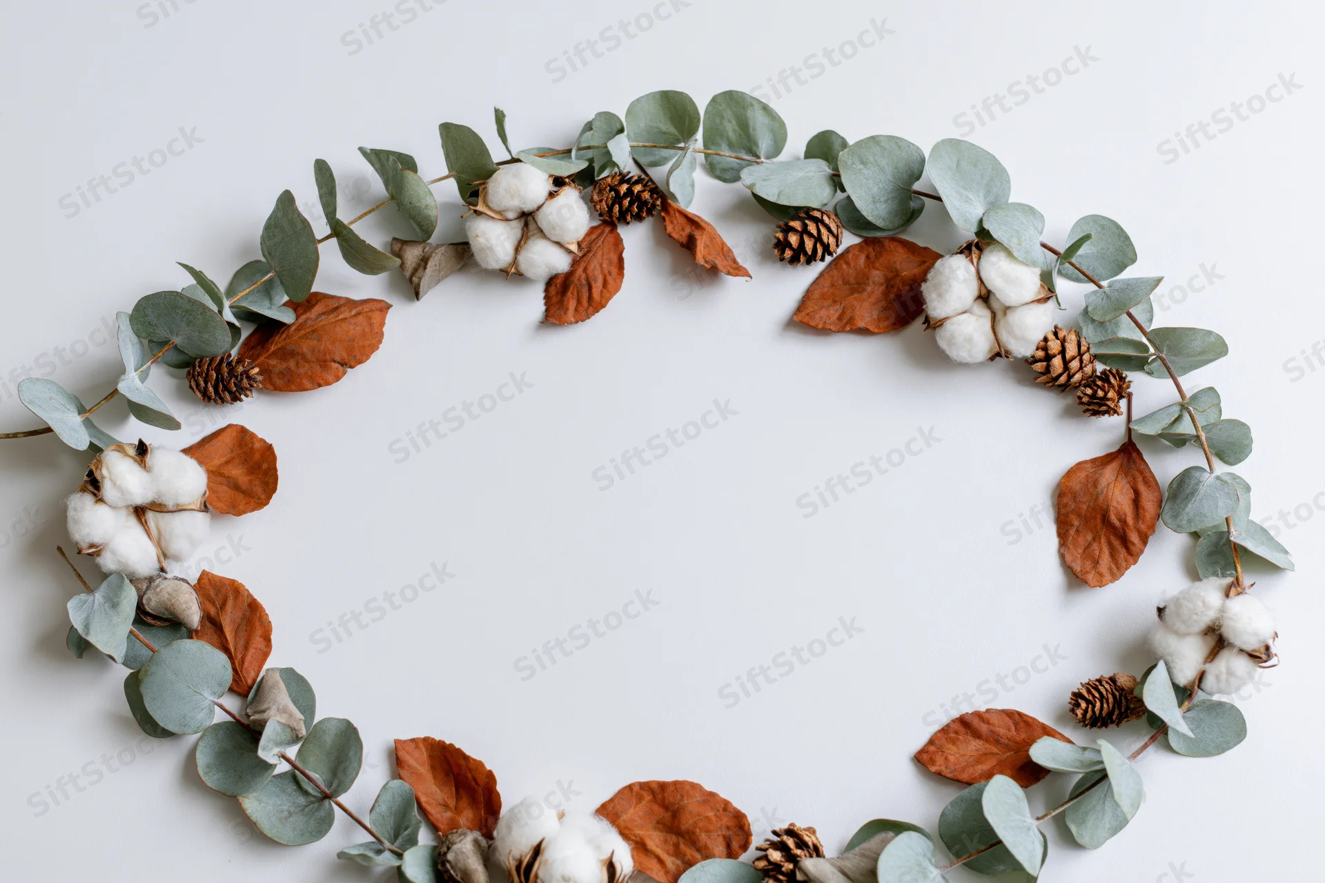 Circular wreath made of eucalyptus branches, dried cotton bolls and pinecones arranged on white surface