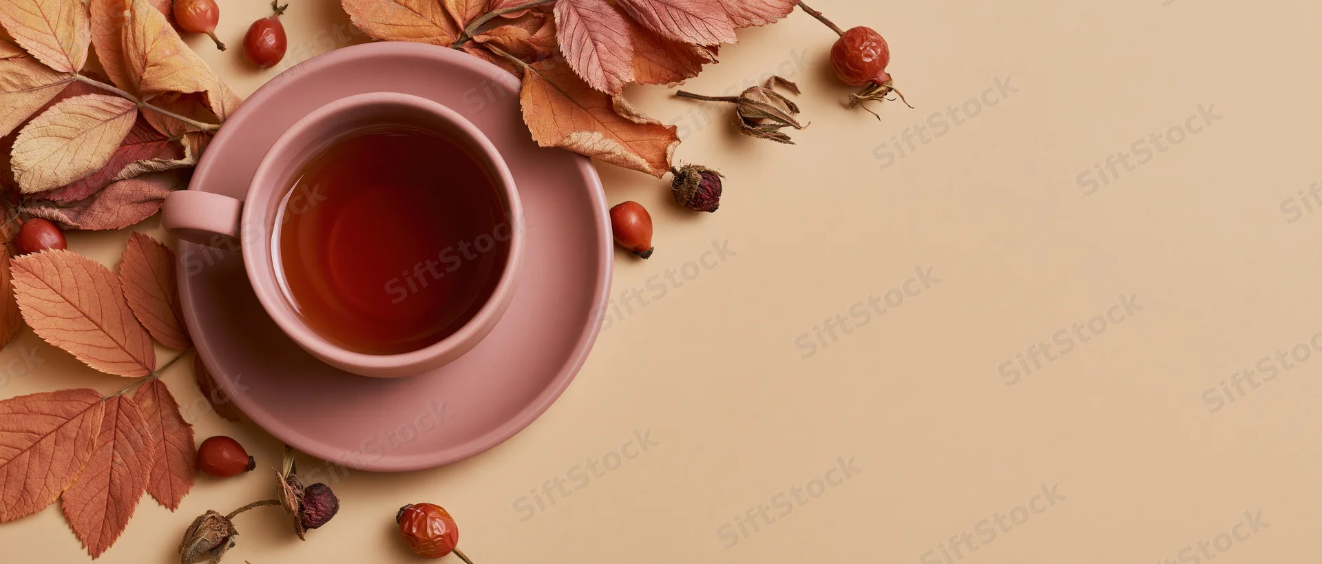 Ceramic cup of tea on pink saucer with autumn leaves and rosehips arranged on beige surface