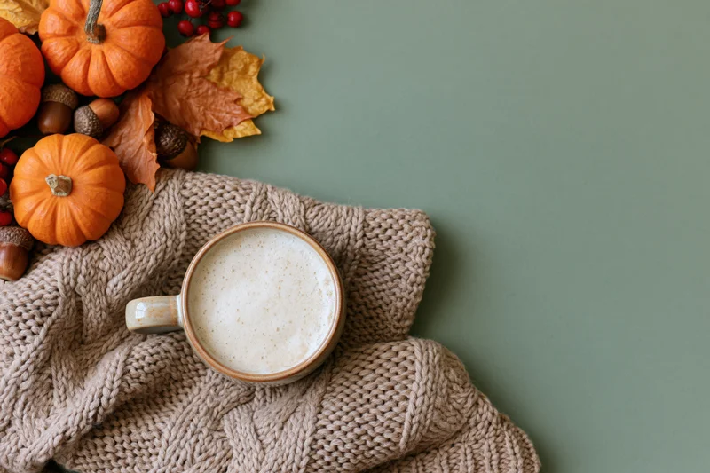 Ceramic mug of latte with foam on a knitted sweater beside autumn decorations on a green surface