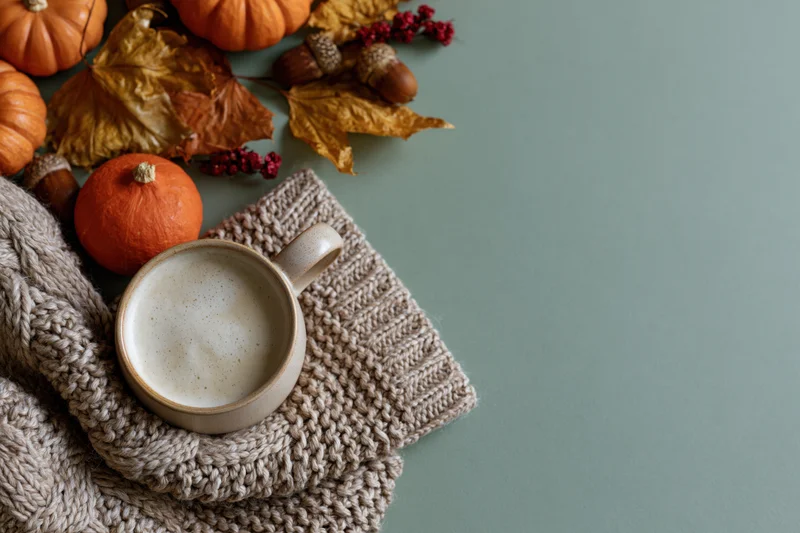 Ceramic mug of latte with foam on knitted sweater and autumn decorations on flat surface
