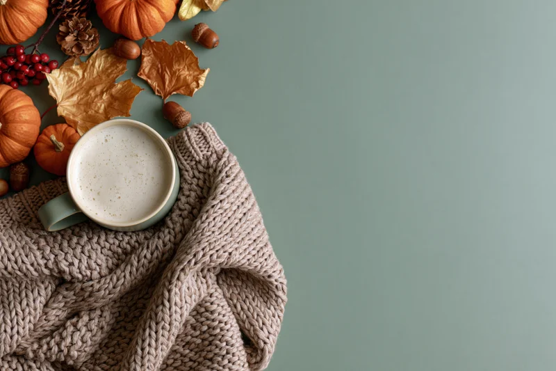 Ceramic mug of latte with foam on a knitted sweater beside autumn decorations on a muted green surface