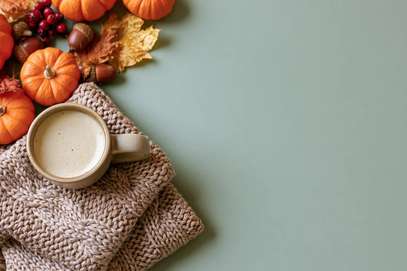 Ceramic mug of latte with foam on knitted sweater beside autumn decorations on green surface