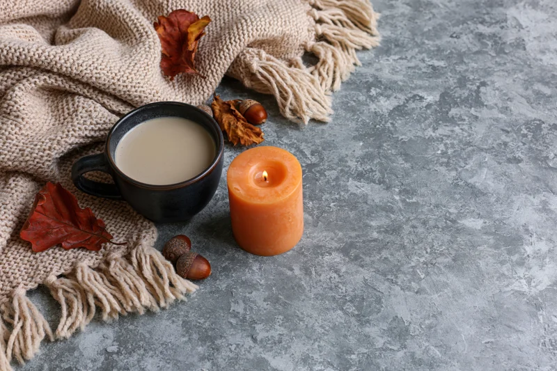Dark ceramic mug with milky coffee, lit orange candle, autumn leaves and acorns on knitted scarf over gray concrete surface