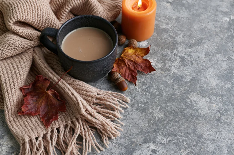 Dark ceramic mug with milky coffee, autumn leaves, acorns, lit orange candle, and beige knit scarf on gray textured surface