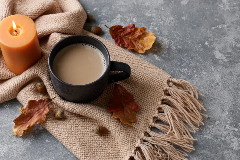 Dark ceramic mug with milky coffee on a knit scarf with autumn leaves and acorns, beside a lit candle on a gray surface