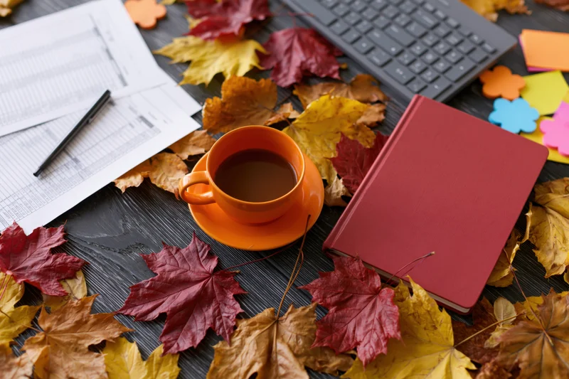 Autumn leaves scattered across a dark wooden desk with office supplies and a cup of tea