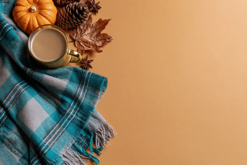 Flat lay of a latte in a gold glass mug with a folded plaid scarf and autumn decorations on a warm-toned surface