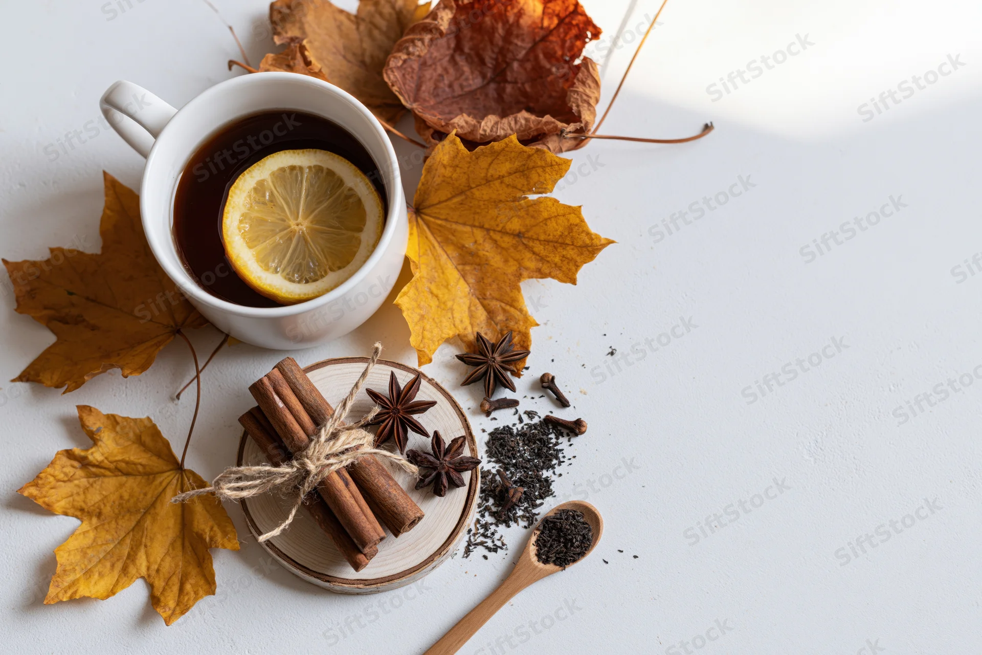 Flat lay of a white ceramic mug with dark tea and lemon slice surrounded by autumn leaves and spices on a light surface