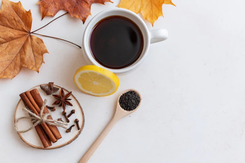 Flat lay of a white ceramic mug filled with dark tea and autumn leaves, cinnamon sticks, lemon slice, and spices on a light surface
