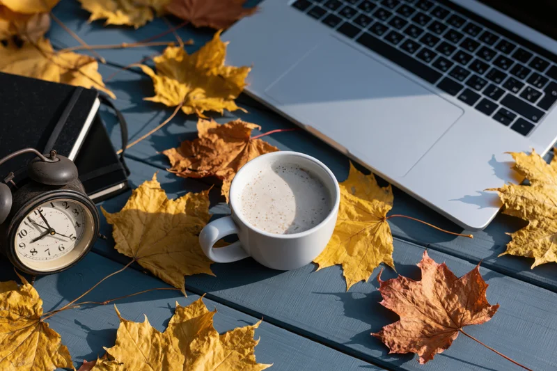 White ceramic mug of milky coffee on blue wooden desk with laptop and autumn leaves in flat lay arrangement