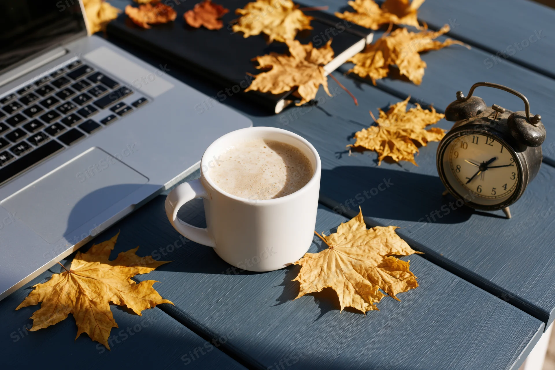 White ceramic mug of milky coffee beside open laptop and vintage alarm clock on blue wooden table scattered with autumn leaves