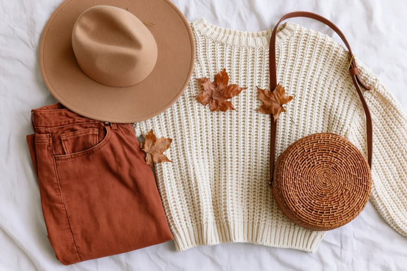 Autumn women's fashion flat lay with sweater, hat, pants, bag and leaves on white background