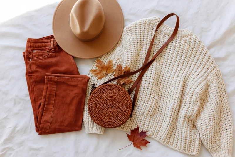 Autumn women's fashion flat lay with sweater, hat, jeans, bag and leaves on white surface