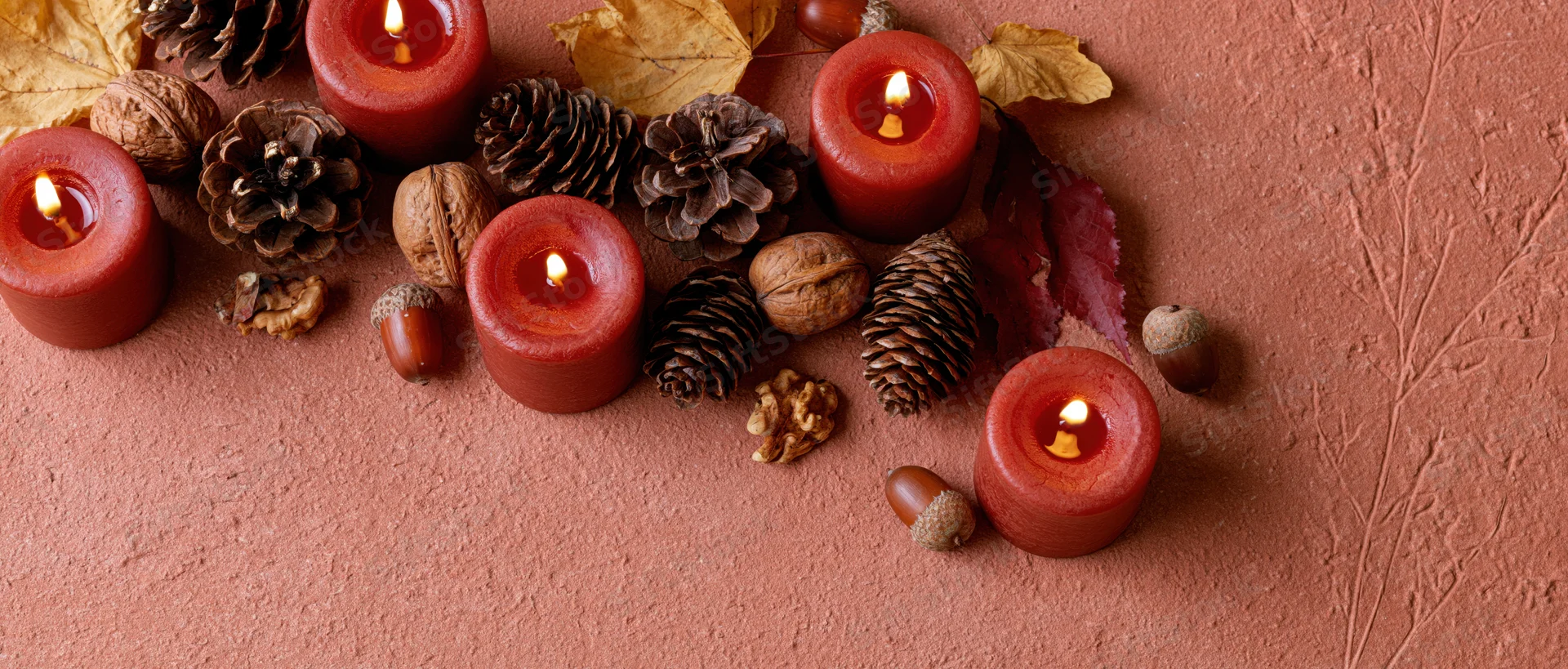 Lit red-orange candles arranged with pinecones, acorns, and autumn leaves on a textured terracotta surface