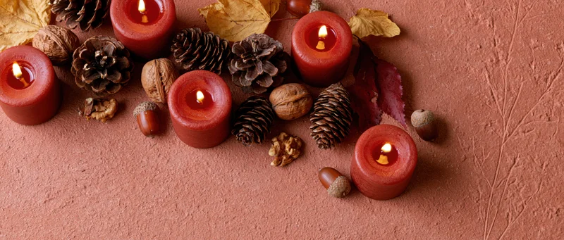 Lit red-orange candles arranged with pinecones, acorns, and autumn leaves on a textured terracotta surface