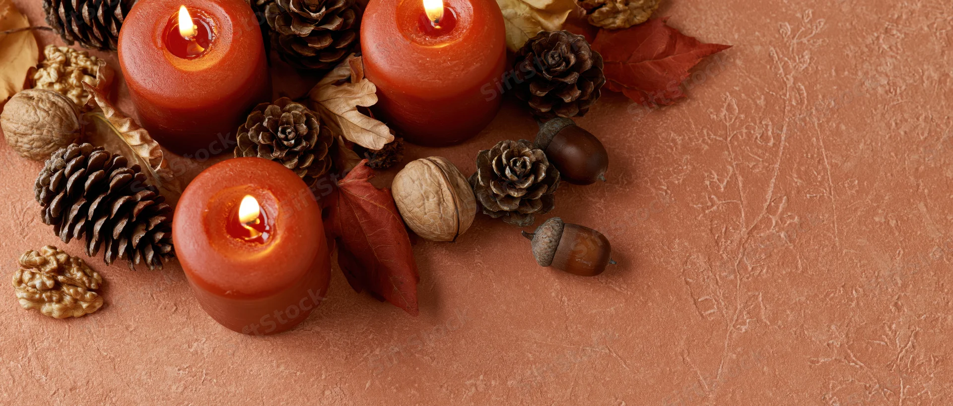 Lit red-orange candles with pinecones, acorns and autumn leaves arranged on textured surface