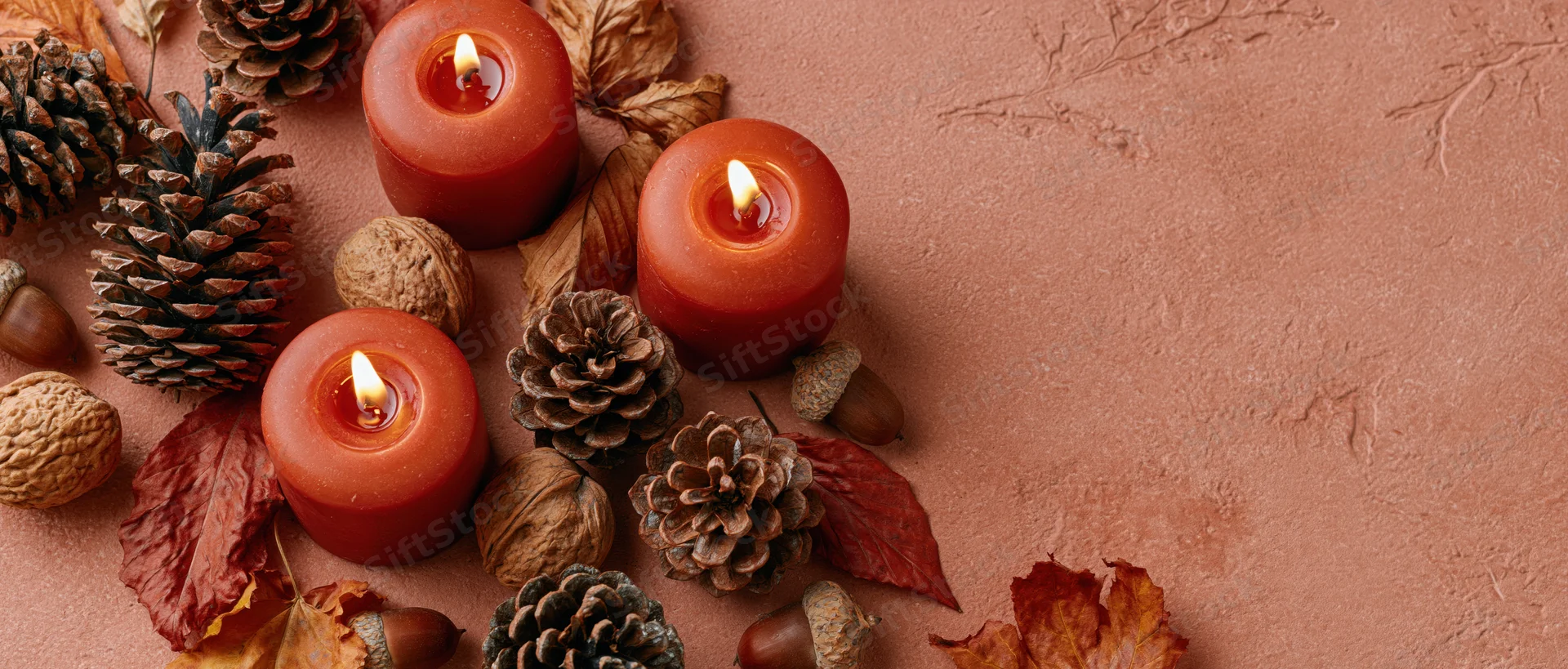 Lit red-orange candles arranged with pinecones, leaves and nuts on a textured terracotta surface