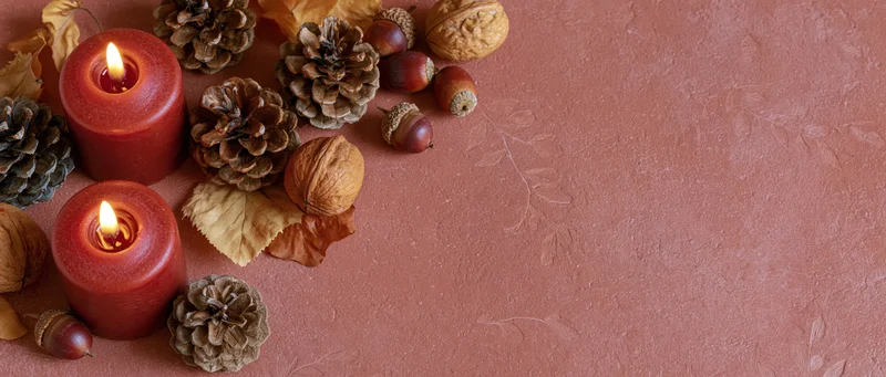 Lit red-orange pillar candles surrounded by pinecones, acorns, walnuts and dried leaves on a textured terracotta surface
