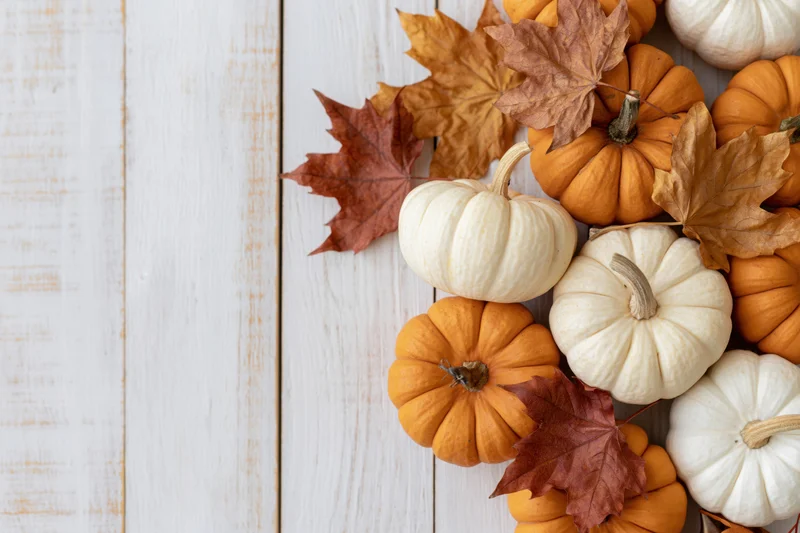 Small orange and white pumpkins with dried maple leaves arranged on a white wooden surface