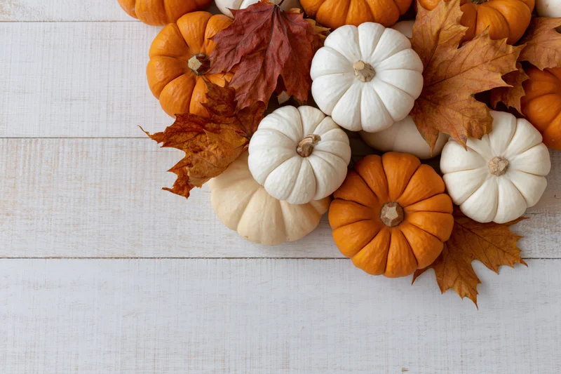 Small orange and white pumpkins arranged with dried autumn leaves on a white wooden table