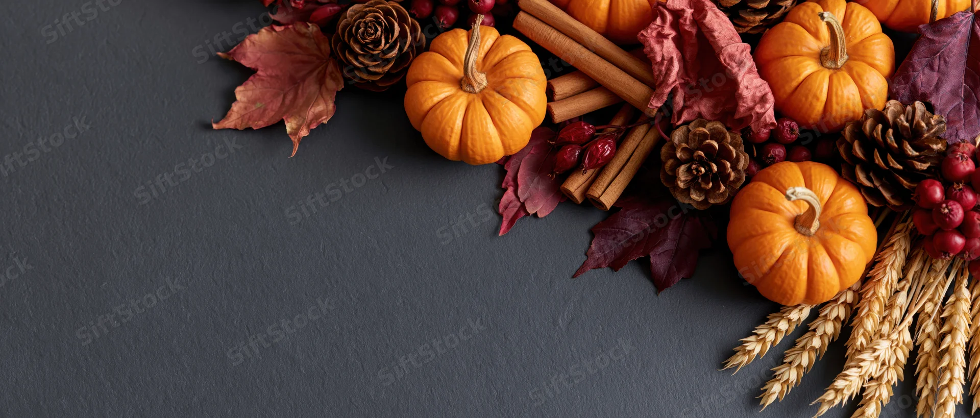 Small orange pumpkins and dried maple leaves arranged with pine cones, cinnamon sticks, berries, and wheat stalks on a dark gray surface