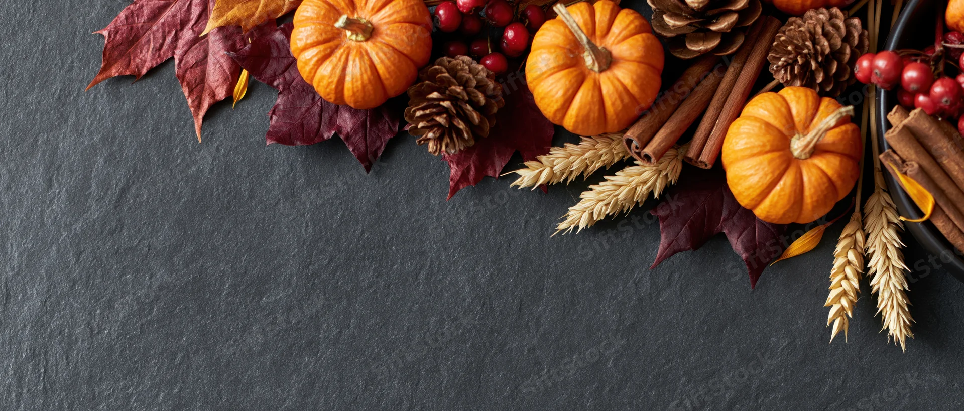 Autumnal flat lay with pumpkins, dried maple leaves, pinecones, cinnamon sticks, wheat, and berries arranged along the top of a dark slate surface