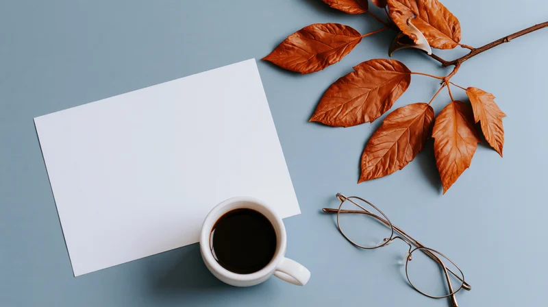 Flat lay overhead view of blank white card, coffee cup, autumn leaves and glasses on blue surface