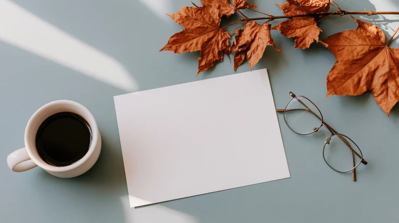 Blank white card with coffee cup, glasses and autumn leaves on blue surface, overhead flat lay