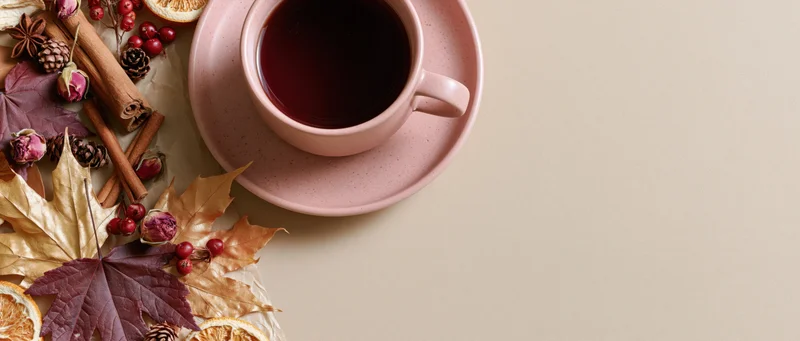 Pink ceramic cup of dark tea on saucer with autumn leaves, cinnamon sticks, pinecones and dried berries arranged on table surface