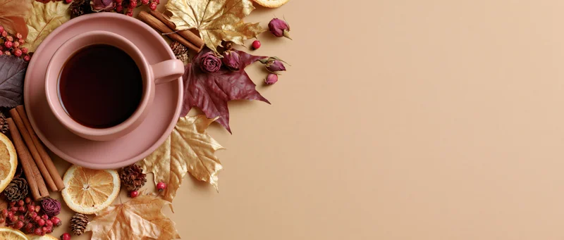 Pink ceramic cup of dark tea surrounded by autumn leaves and spices on a flat surface with copyspace