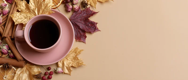 Pink ceramic cup of dark tea surrounded by autumn leaves and spices on beige surface