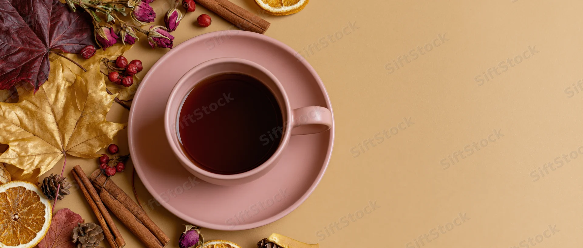 Pink ceramic cup of dark tea on saucer surrounded by autumn leaves, cinnamon sticks, dried oranges and berries on beige surface