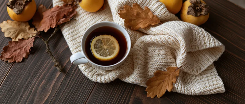 White ceramic mug with lemon tea on knitted sweater and autumn leaves on wooden table