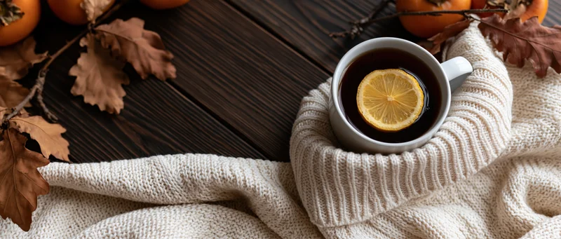 White ceramic mug with lemon tea resting on a knitted sweater on dark wooden surface surrounded by autumn leaves and pumpkins
