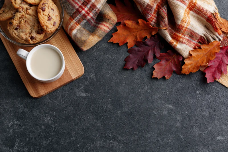 White ceramic mug of milky coffee on wooden tray with chocolate chip cookies, autumn leaves and plaid scarf on dark surface
