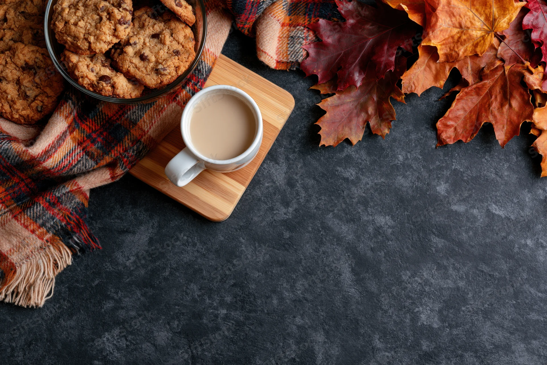 White ceramic mug of milky coffee on wooden tray with cookies, plaid scarf and autumn leaves on dark surface