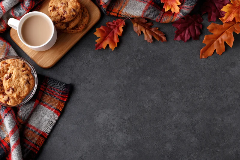 White ceramic mug of milky coffee with chocolate chip cookies and autumn leaves on a dark surface with plaid scarf and wooden tray, flat lay overhead view