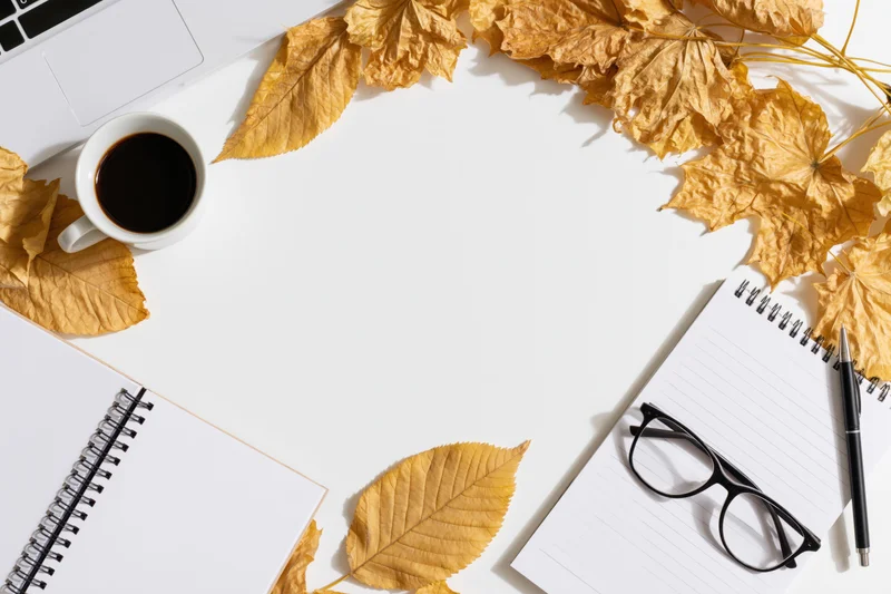 Dried autumn leaves arranged around a white desk with laptop, notebook, coffee cup, pen and glasses, flat lay overhead view