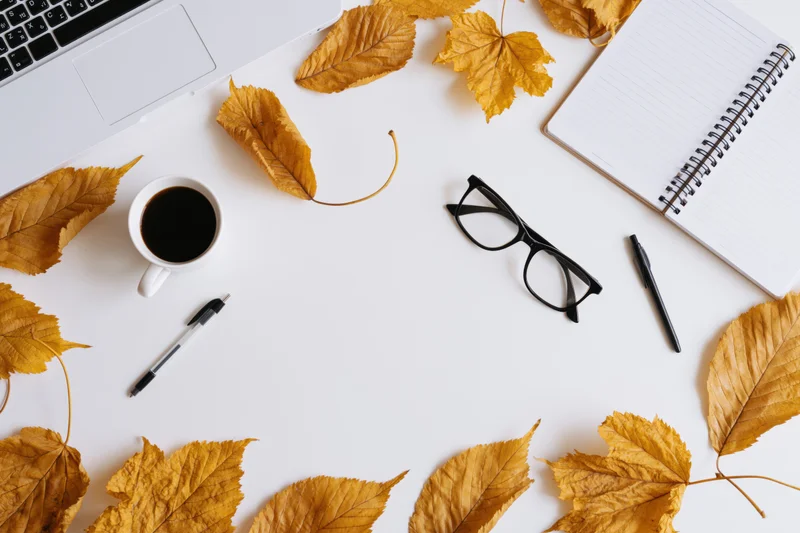 Overhead view of a white desk with laptop, notebook, coffee cup, glasses, pens and autumn leaves arranged around the edges