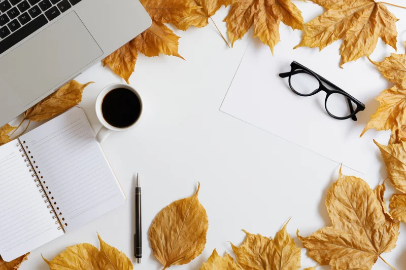 Autumn leaves arranged around a laptop, notebook, coffee cup and glasses on a white desk surface