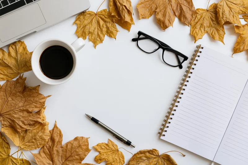 Autumn leaves arranged around a laptop, coffee cup, notebook and glasses on a white desk surface