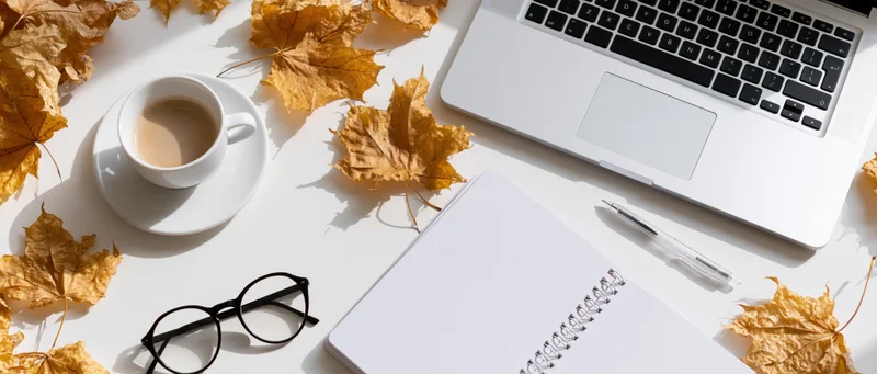 Overhead view of a white desk with a laptop, coffee cup, notebook, glasses and autumn leaves scattered around