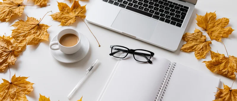 Overhead view of a laptop, notebook, coffee cup and autumn leaves arranged on a white desk surface