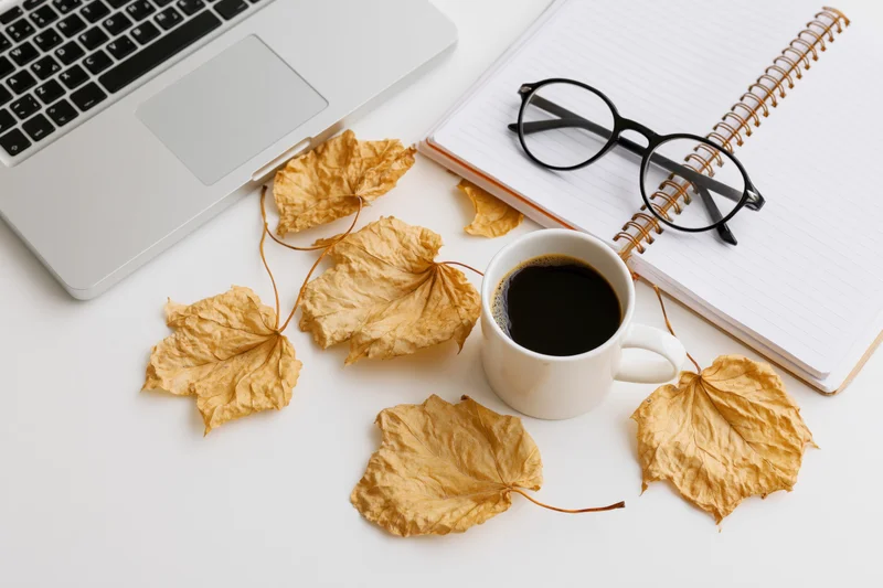 Overhead view of a white desk with a laptop, coffee cup, notebook, glasses and dried autumn leaves arranged for work or study