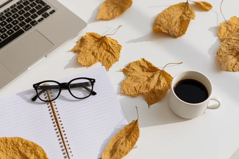 Overhead view of a white desk with a laptop, notebook, glasses, coffee cup and dried autumn leaves arranged for a workspace setup
