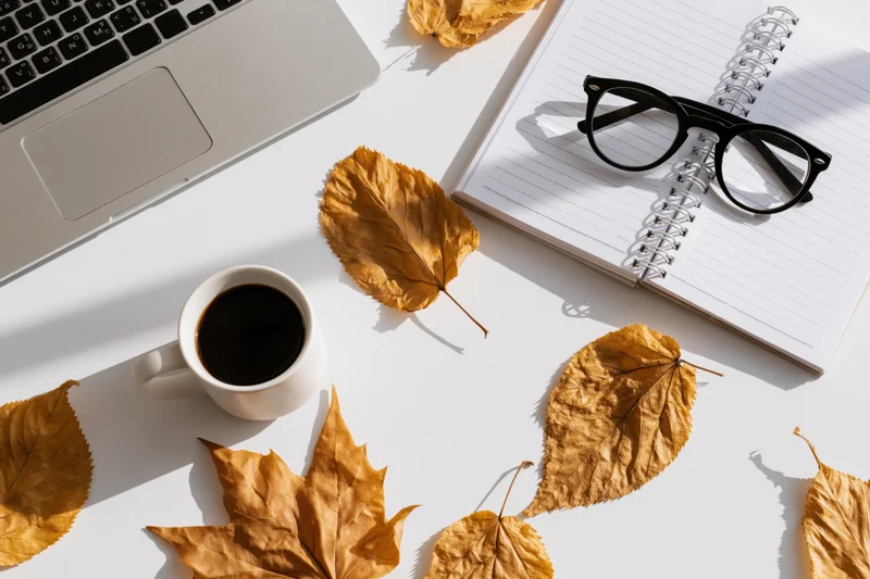Overhead view of a white desk with laptop, notebook, coffee mug, and autumn leaves arranged for work or study