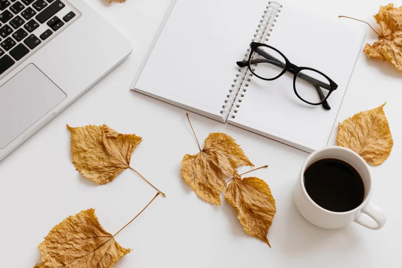 Overhead view of a white desk with a laptop, open notebook, coffee cup, glasses and dried autumn leaves arranged for work or study