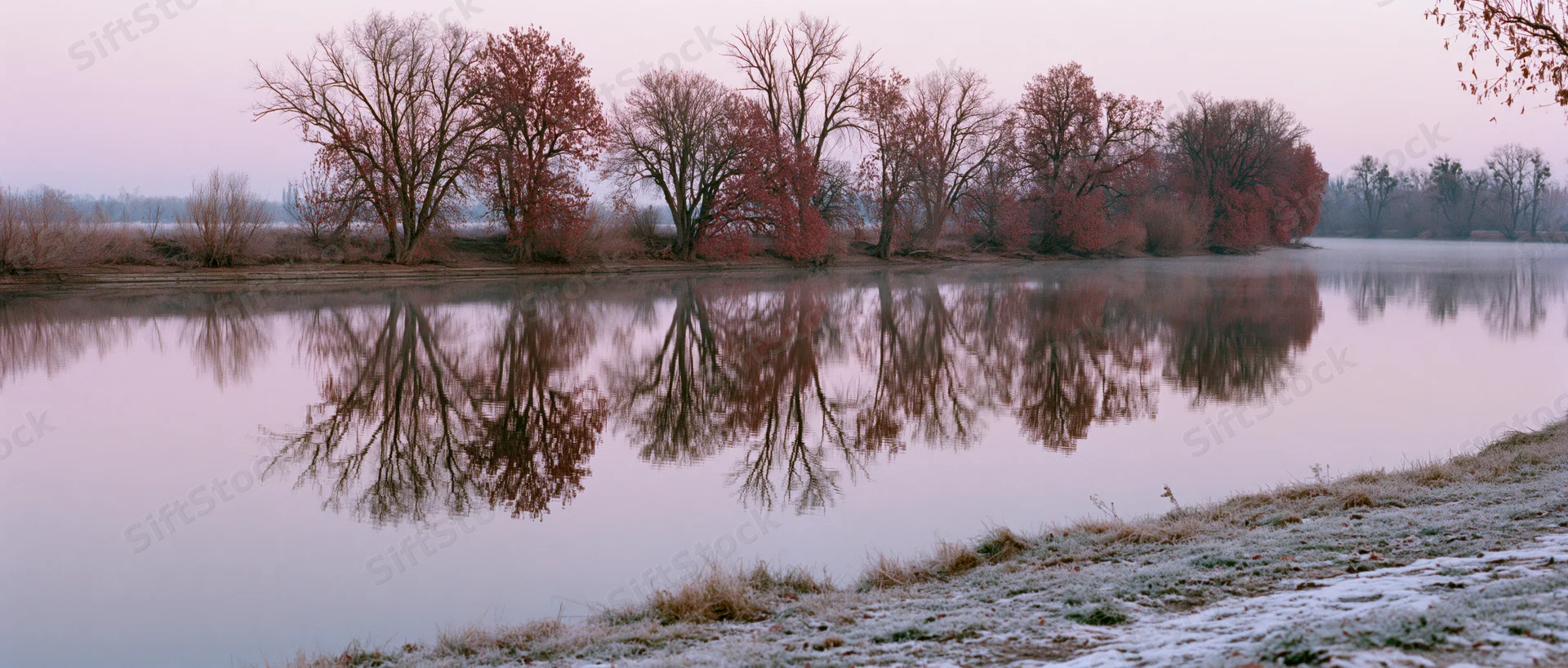 Frost-covered riverbank at sunrise with bare trees reflected in calm water