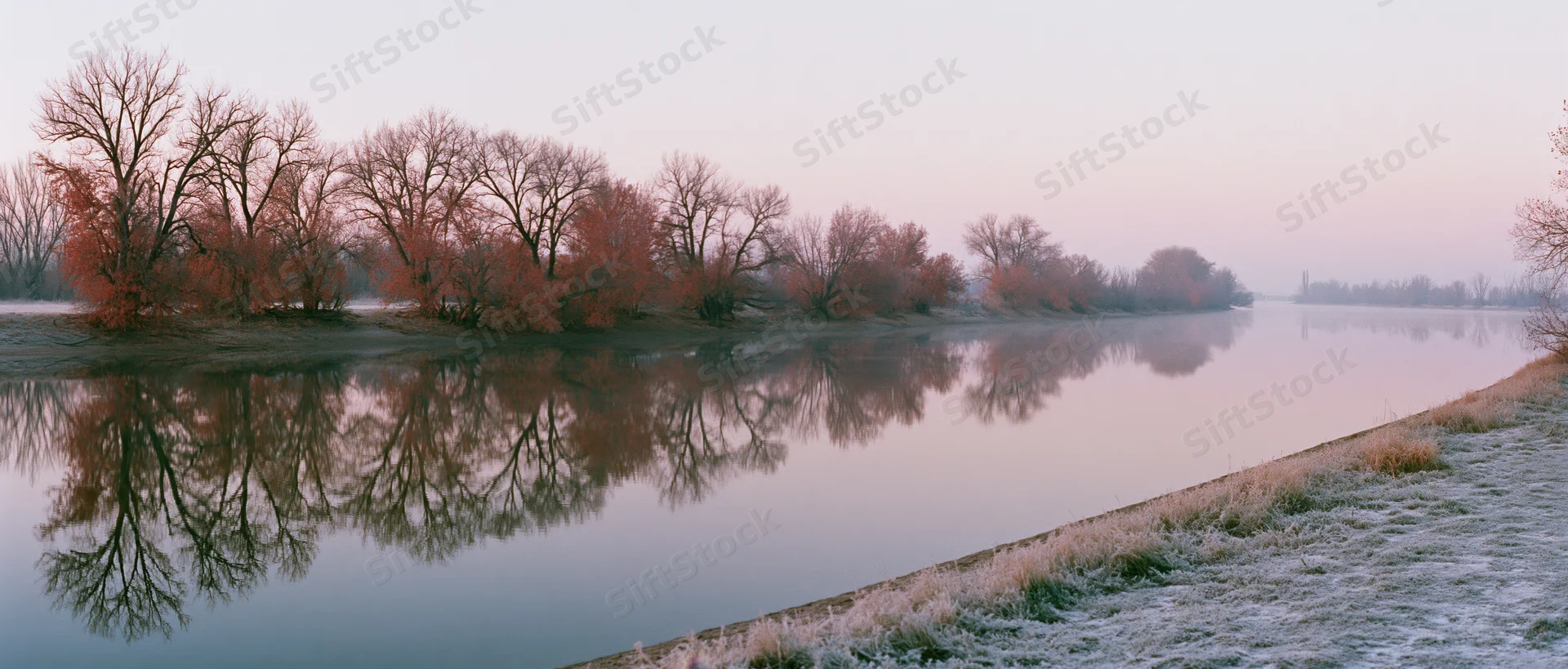 Frost-covered riverbank at sunrise with bare trees reflected in calm water